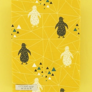 May include: Yellow fabric featuring a penguin pattern in black and white, geometric lines, and teal, black, and white triangles. The fabric includes the text "OTTOBRE design" on a label. A ruler is also visible.