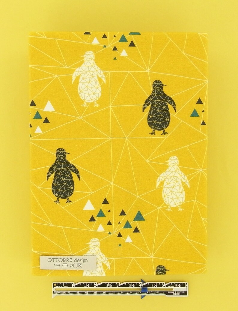 May include: Yellow fabric featuring a penguin pattern in black and white, geometric lines, and teal, black, and white triangles. The fabric includes the text "OTTOBRE design" on a label. A ruler is also visible.