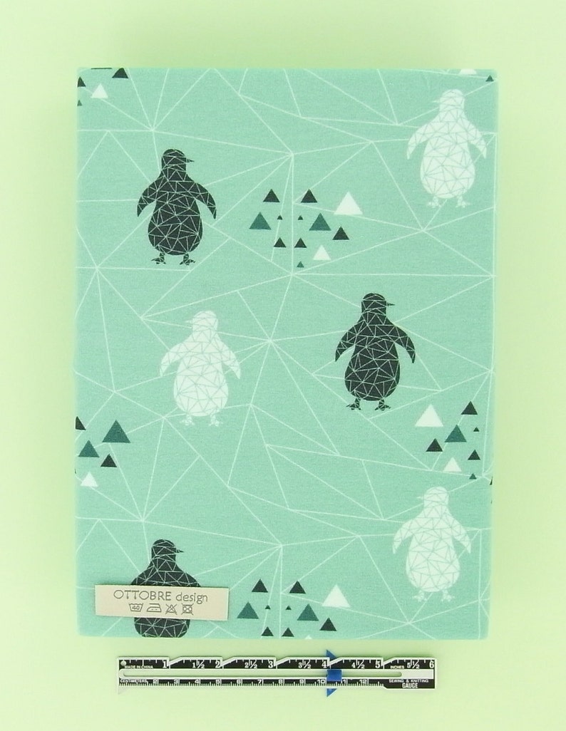 May include: A rectangular fabric piece with a light teal background featuring a geometric pattern of white lines, black and white penguins, and triangles. A small label reads "OTTOBRE design". A ruler is at the bottom.