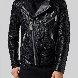 Könnte beinhalten: Eine schwarze Jacke mit einer strukturierten, schlangenlederartigen Oberfläche, ausgestattet mit einem diagonalen Frontreißverschluss, einem Stehkragen mit Druckknopf sowie mehreren silberfarbenen Reißverschlusstaschen an Brust und Seiten.