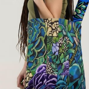 May include: A tote bag with a vibrant botanical design featuring blue, green, and purple succulents. The bag has black straps and a matching rolled-up item inside. The artwork is detailed and colorful, creating a striking visual appeal.