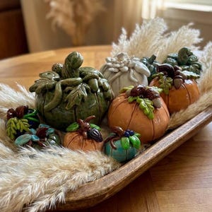 May include: A collection of small ceramic pumpkins in various colours including green, orange, white, and blue, each decorated with sculpted leaves and acorns, arranged inside a long wooden bowl filled with dried pampas grass plumes.