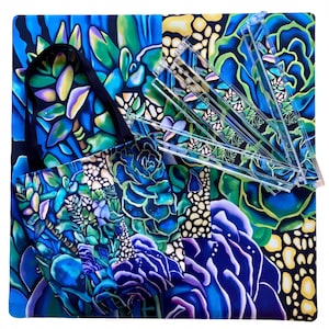 May include: A tote bag and several clear plastic rulers rest on a square, patterned surface. The tote bag and surface feature a vibrant, colorful design of blue, green, and purple succulents. The tote bag has a black handle.