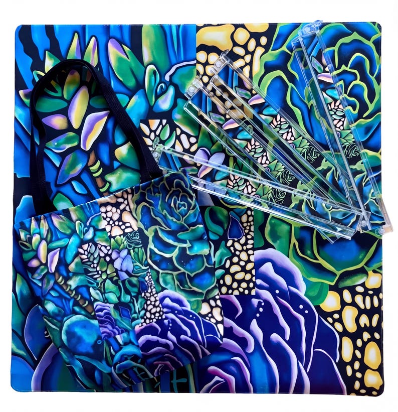 May include: A tote bag and several clear plastic rulers rest on a square, patterned surface. The tote bag and surface feature a vibrant, colorful design of blue, green, and purple succulents. The tote bag has a black handle.