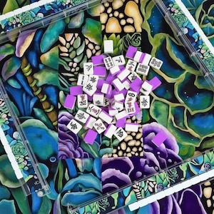 May include: A colorful Mahjong set with white tiles featuring black Chinese characters and purple backs. The tiles are scattered on a vibrant floral patterned surface, framed by clear plastic trays.