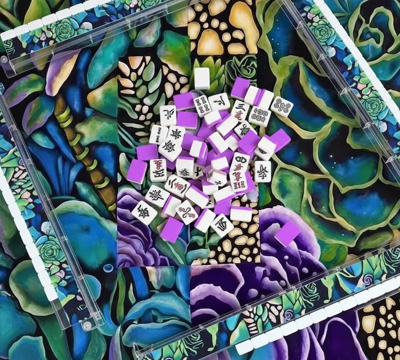 May include: A colorful Mahjong set with white tiles featuring black Chinese characters and purple backs. The tiles are scattered on a vibrant floral patterned surface, framed by clear plastic trays.