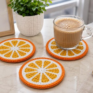 Puede incluir: Tres posavasos redondos de ganchillo con diseño de rodaja de naranja, con segmentos amarillos, borde blanco y contorno naranja. Un posavasos sostiene una taza de cristal con una bebida marrón claro, mientras otros dos están planos.