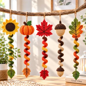 Könnte beinhalten: Fünf gehäkelte Hängedekorationen, die an einem Ast befestigt sind. Das Set besteht aus einer Sonnenblume, einem Kürbis, einem Ahornblatt, einer Eichel und einem Eichenblatt. Jedes Ornament hat einen langen, bunten, spiralförmigen Schwanz.