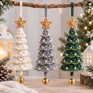 May include: Three crocheted Christmas tree ornaments in white, gray, and dark green. Each tree features a gold star topper, small white pearl accents, gold thread trim, and a gold jingle bell hanging from the bottom point of the tree.