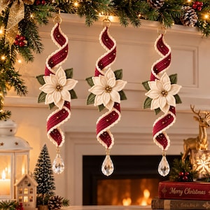 Könnte beinhalten: Drei hängende Ornamente mit einem spiralförmigen, dunkelroten Band mit cremefarbenen Rändern, akzentuiert durch eine zentrale weiße Weihnachtssternblüte mit grünen Blättern und Goldperlen, abgeschlossen mit einem klaren, tropfenförmigen Kristall.