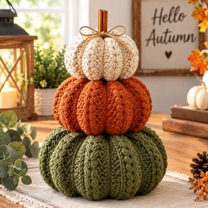May include: A stack of three crocheted pumpkins in cream, orange, and olive green. The pumpkins feature a cable knit texture and are topped with a cinnamon stick stem tied with a thin jute twine bow. The stack is arranged vertically.