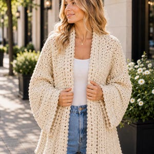 May include: A cream-colored, open-front crochet cardigan featuring wide, loose sleeves and a textured, scalloped edge along the front opening. The garment is worn over a simple white top and light blue denim jeans, showcasing a chunky knit pattern.