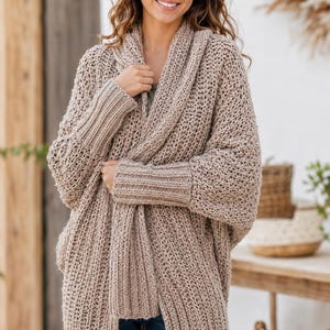 Könnte beinhalten: Eine beigefarbene, vorne offene Strickjacke mit grober Strickstruktur, einem breiten Schalkragen und langen Ärmeln mit gerippten Bündchen. Das Kleidungsstück hat eine lockere, übergroße Passform und wird über einer blauen Jeans getragen.