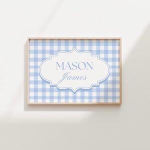 May include: A framed print featuring a light blue and white gingham pattern background. A white and light blue ornate oval shape in the center displays the name "MASON" above "James" in a script font. The frame is a light wood color.
