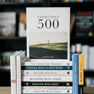 May include: A stack of books with the title "The Faith Vault 500" on top, featuring a cross on a hill. Other titles include "The Power of Prayer" and "Walking with Jesus". The books are on a wooden surface.
