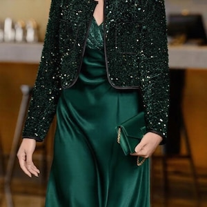 Pode incluir: Um conjunto verde esmeralda escuro composto por um vestido longo de cetim e um casaco curto a condizer. O casaco apresenta adornos de lantejoulas densas e acabamentos pretos. Uma pequena bolsa de mão de cetim a condizer é segurada na mão.