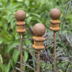 Può includere: Tre picchetti da giardino in metallo con un terminale sferico su una base quadrata con una finitura marrone ruggine testurizzata. Ogni picchetto è costituito da una sottile asta verticale che sostiene l'ornamento, con un aspetto in ferro ossidato.