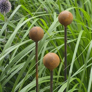 Op de afbeelding: Drie bolvormige tuinornamenten met een verweerde, roestbruine metalen afwerking, elk gemonteerd op een dunne, bijpassende metalen staaf. De bollen zijn op verschillende hoogtes geplaatst, wat een verticale opstelling vormt tussen het gras.