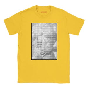 Puede incluir: Una camiseta amarilla de manga corta con un estampado gráfico rectangular en blanco y negro en el pecho. El gráfico consiste en un patrón denso de pequeños caracteres de texto que forman una imagen. El texto MENOS STUDIO aparece en rojo en el cuello.