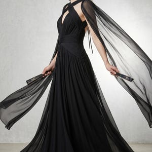 May include: A black evening gown with a flowing skirt and a halter neckline. The dress features a cape that drapes over the shoulders. The model is holding a fan. The dress is made of a lightweight, sheer fabric.