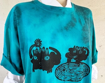 May include: A teal tie-dye t-shirt featuring a black graphic print of two stylized creatures, one standing near a circular patterned pool. The shirt is layered over a white collared shirt on a mannequin with the text MUMIAI visible on the neck.