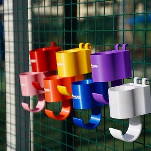 Puede incluir: Colección de coloridos portavasos de plástico en rojo, rosa, naranja, amarillo, azul, morado y blanco, sujetos a una valla de alambre verde. Cada soporte tiene un gancho.