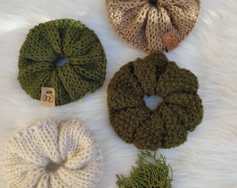 May include: Four knitted hair scrunchies in different colors: green, brown, beige, and white. The scrunchies are all handmade and have a small leather tag with a bow design.
