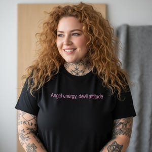 May include: Black t-shirt with the text "Angel energy, devil attitude" in pink. The shirt is a crew neck style. The person wearing the shirt has tattoos on their arms and neck.