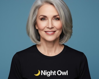 以下が含まれることがあります： 灰色の髪の女性が笑顔で、黒いTシャツを着ています。「Night Owl」の文字と黄色の三日月ロゴが入っています。背景は単色の青色です。