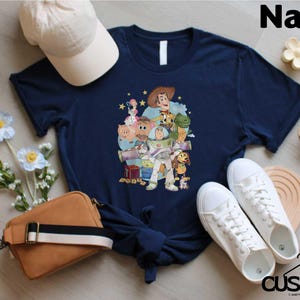 A navy blue t-shirt with a graphic of characters from Toy Story, including Woody, Buzz Lightyear, and other beloved characters, along with a baseball cap, a brown purse, and a pair of white sneakers.