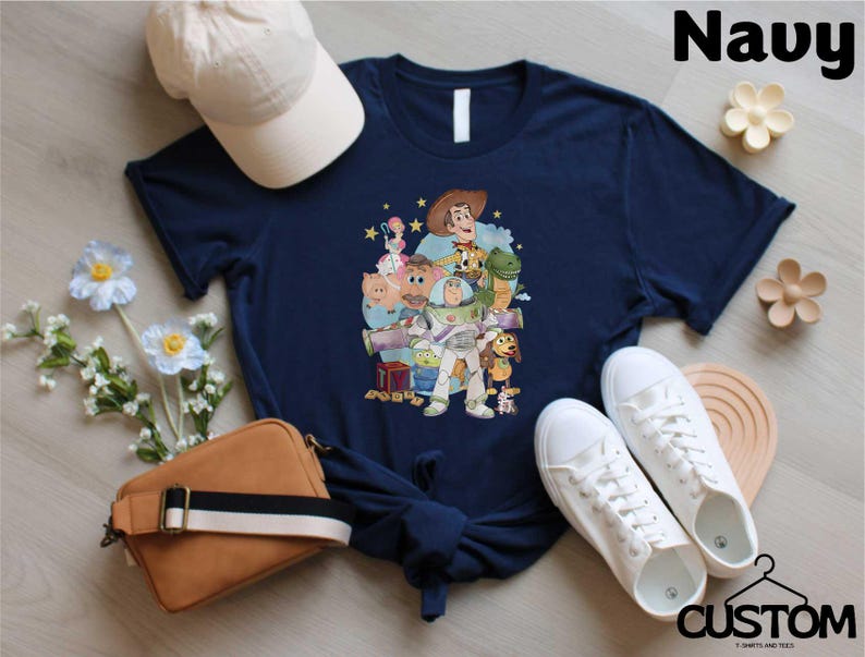 A navy blue t-shirt with a graphic of characters from Toy Story, including Woody, Buzz Lightyear, and other beloved characters, along with a baseball cap, a brown purse, and a pair of white sneakers.