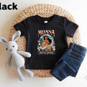 A black t-shirt with a graphic of Moana, a character from the Disney animated film "Moana", along with a wicker basket and a crocheted rabbit toy.