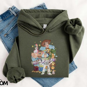 A green hoodie with a cartoon character design, along with a pair of blue jeans, laid out on a white surface.