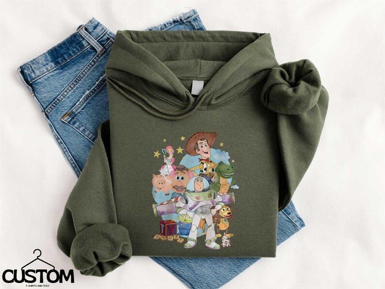 A green hoodie with a cartoon character design, along with a pair of blue jeans, laid out on a white surface.