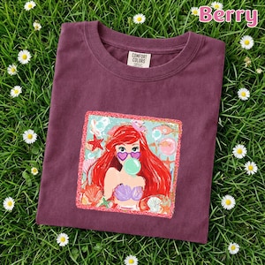 May include: A berry-colored crew neck t-shirt featuring a square graphic print of a red-haired mermaid wearing heart-shaped sunglasses and blowing a bubble. The print includes seashells, starfish, and bubbles on a light blue background.