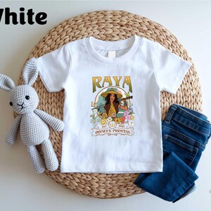 A white t-shirt with a graphic design featuring a woman with a guitar, surrounded by flowers and text, placed on a woven placemat. Next to the t-shirt is a crocheted stuffed rabbit, and a pair of blue jeans.