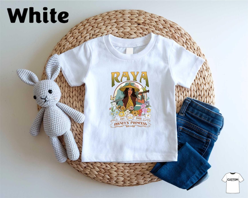 A white t-shirt with a graphic design featuring a woman with a guitar, surrounded by flowers and text, placed on a woven placemat. Next to the t-shirt is a crocheted stuffed rabbit, and a pair of blue jeans.