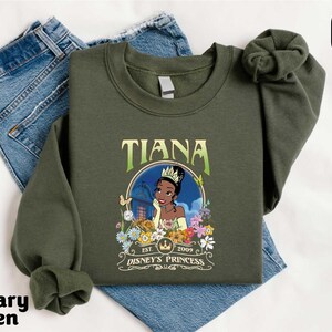 A green sweatshirt with a graphic of a character from the Disney animated film " princess" printed on it, along with the text "Est. 2005" and "Disney's Princess". The sweatshirt is placed on top of a pair of blue jeans.