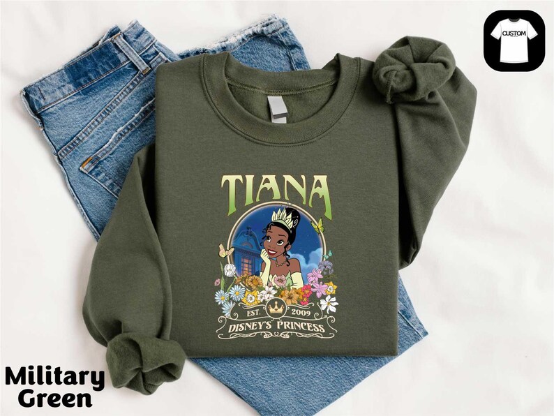 A green sweatshirt with a graphic of a character from the Disney animated film " princess" printed on it, along with the text "Est. 2005" and "Disney's Princess". The sweatshirt is placed on top of a pair of blue jeans.