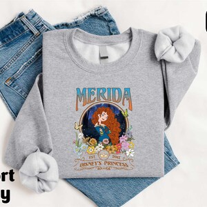 A gray sports t-shirt with a graphic design featuring Merida, the protagonist from the Disney animated film "Disney Princess", along with the text "Est. 2012" and "Disney Princess". The t-shirt is placed on top of a pair of blue jeans.