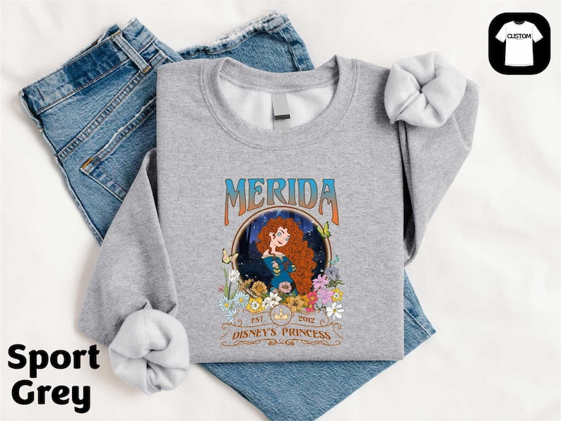 A gray sports t-shirt with a graphic design featuring Merida, the protagonist from the Disney animated film "Disney Princess", along with the text "Est. 2012" and "Disney Princess". The t-shirt is placed on top of a pair of blue jeans.