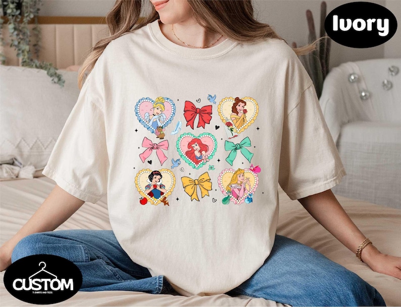 a woman sitting on a bed wearing a t - shirt with princesses on it