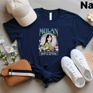 A navy blue t-shirt with a graphic of Mulan, a character from the Disney animated film "Mulan", along with a white baseball cap, a brown purse, and white sneakers. The t-shirt is displayed on a wooden surface, surrounded by various accessories and flowers.