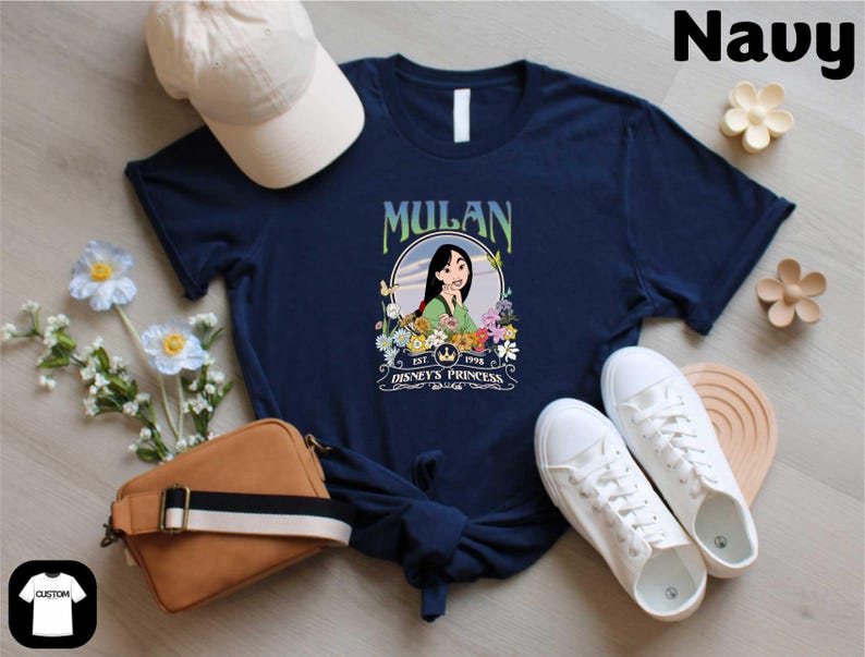 A navy blue t-shirt with a graphic of Mulan, a character from the Disney animated film "Mulan", along with a white baseball cap, a brown purse, and white sneakers. The t-shirt is displayed on a wooden surface, surrounded by various accessories and flowers.