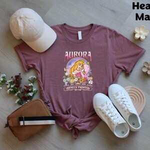 a purple t-shirt with a graphic of a character from the Disney movie "Aurora", along with a white baseball cap, a brown purse, a pair of white sneakers, and some flowers.