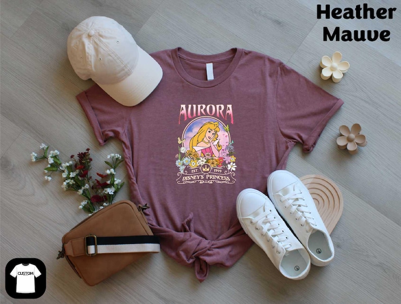 a purple t-shirt with a graphic of a character from the Disney movie "Aurora", along with a white baseball cap, a brown purse, a pair of white sneakers, and some flowers.