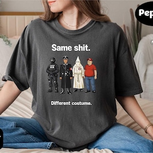 May include: Dark gray t-shirt with the text "Same shit. Different costume." and illustrations of four figures in different uniforms. The shirt is a relaxed fit and has short sleeves.