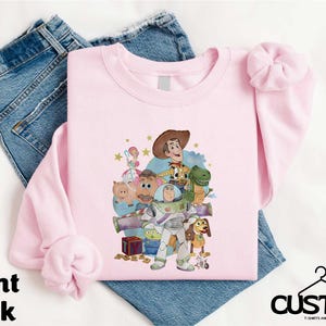 A pink t-shirt with a cartoon character design, along with a pair of blue jeans, laid out on a white background.