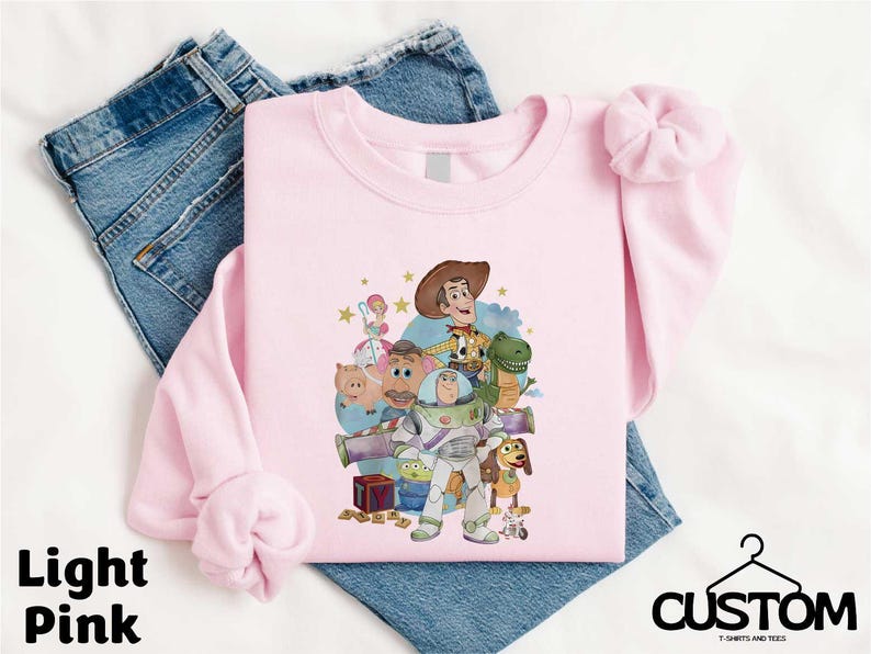 A pink t-shirt with a cartoon character design, along with a pair of blue jeans, laid out on a white background.