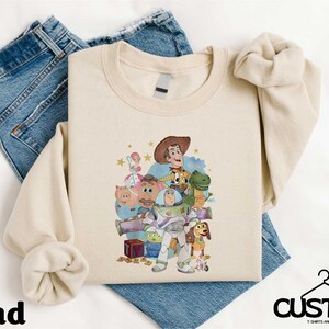 A beige t-shirt with a cartoon character design, including a cowboy hat, a dinosaur, and other characters, is displayed on a white background.